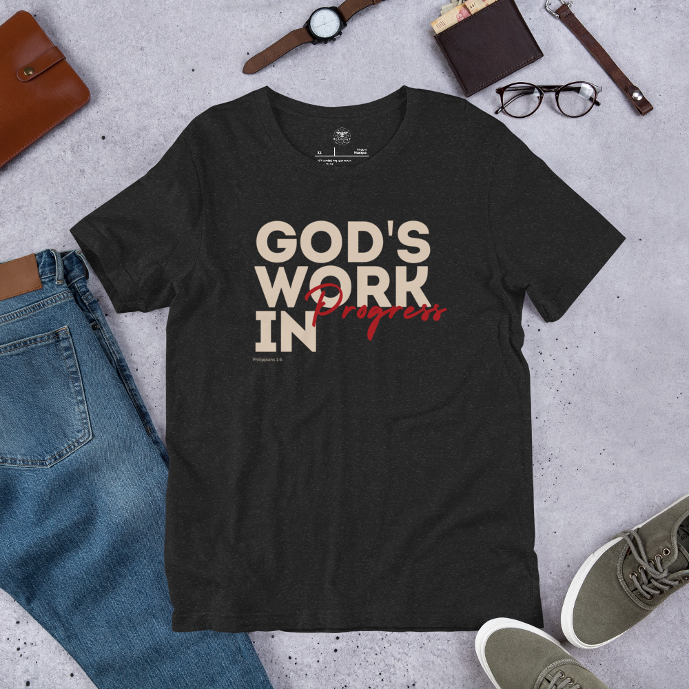 Front view of Heavenly Crafted's 'God's Work in Progress' T-shirt in Charcoal Black with bold white text. Inspired by Philippians 1:6, it's a reminder of God's ongoing work in your life. Laid out on the floor with glasses a wallet a watch a pair of shohes and so blue genes