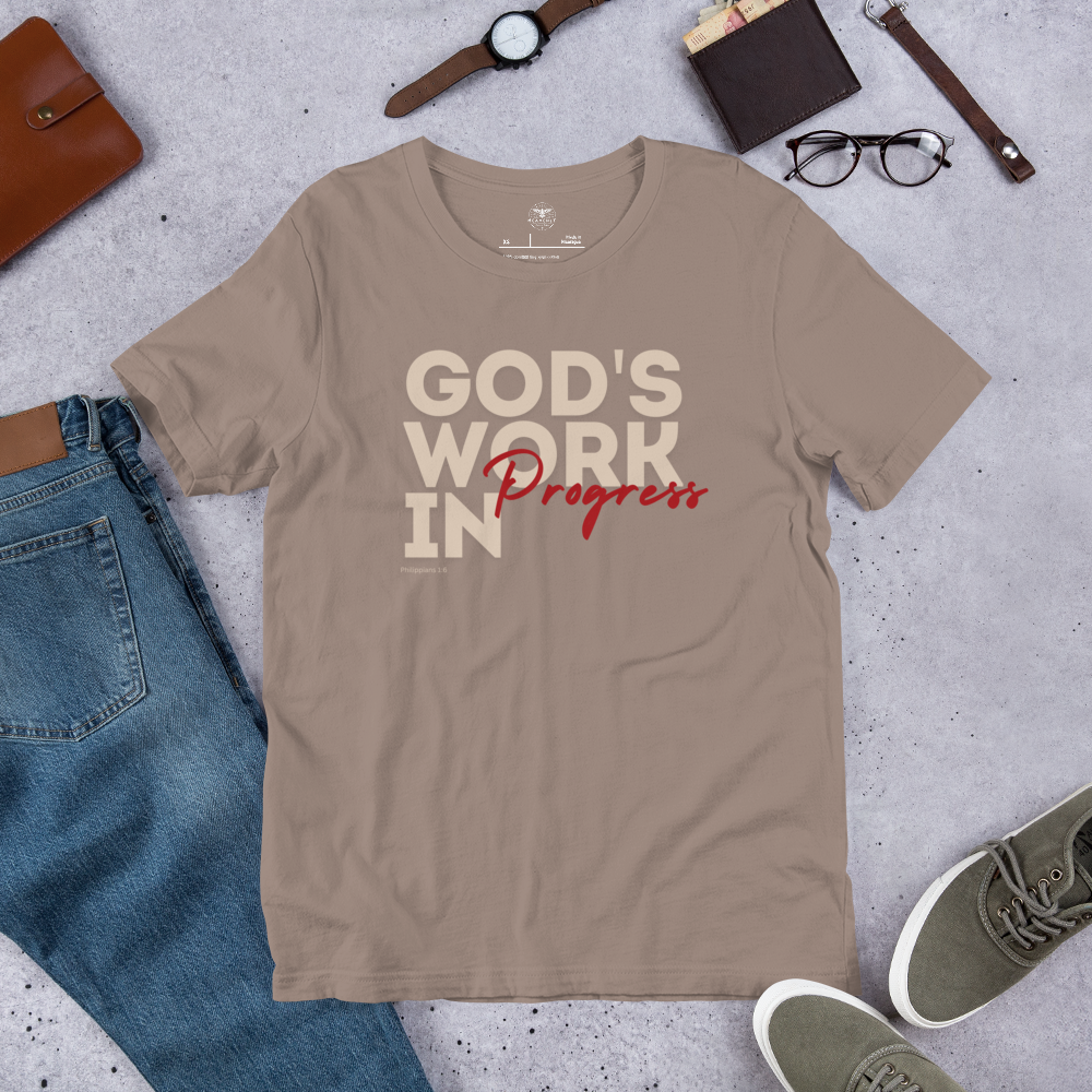 Front view of Heavenly Crafted's 'God's Work in Progress' T-shirt in Tan, showcasing the inspirational text design in bold lettering. A reminder of faith and growth inspired by Philippians 1:6. Laid out on the floor with glasses, a wallet, a watch, pair of shoes, blue genes, and a journal