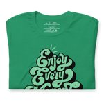 Main display mockup of 'Enjoy Every Moment' T-shirt in a relaxed store setting, featuring its bold graffiti lettering on a vibrant green tee. Ideal for adding a touch of inspiration to any wardrobe.