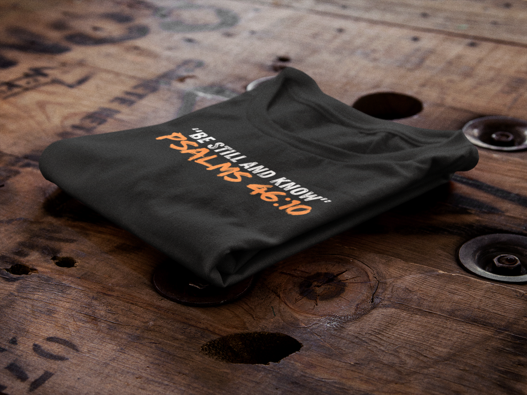 Black 'Be Still and Know' T-shirt, inspired by Psalm 46:10, folded neatly on a wooden spool. Made from 100% cotton, this classic men's tee offers comfort and a structured look. Its durable fabric and thoughtful design details make it perfect for casual wear or layering, reminding you of God's calming presence.